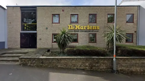 Google Light-brown brick-built two-storey factory with small square windows and yellow Dr Marten's logo on the front of the building. There are steps to the front entrance and there are two palm trees in the foreground.