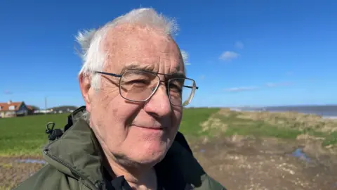 Amanda White / BBC A head and shoulders shot of a man smiling at the camera. He has short grey hair and is wearing large glasses and a green waterproof coat. He is standing on a path on a cliff near the coast and green fields can be seen behind him. The sky is blue. 
