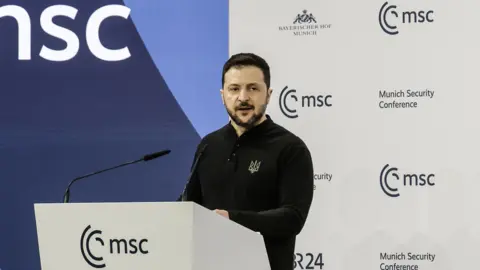 EPA-EFE/REX/Shutterstock Volodymyr Zelensky stands at a lectern at the Munich Security Conference as he gives a speech. He is wearing a black fleece with the symbol of the Ukrainian Defence Forces embroidered on the breast.