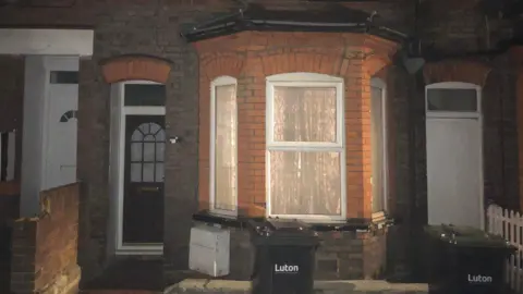 Tony Fisher/BBC The exterior of the ground floor of a redbrick terraced house, which has a bay window and the curtains drawn, with the light on inside. The front door is to the left and outside are two wheelie bins, with the word "Luton" on the front.