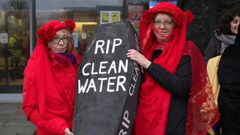 Dua pengunjuk rasa mengenakan pakaian berwarna merah dan memegang peti mati bertuliskan RIP air bersih.