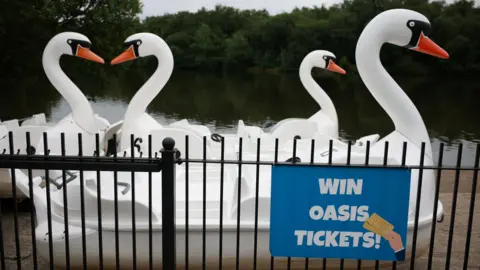 Reuters Four large swan-like pedal boats on a Heaton Park lake behind railings with a blue sign offering the chance to win Oasis concert tickets.