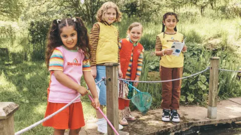 Emily Stein/Girlguiding Four young girls, two Rainbows and two Brownies, standing by a pond in their new uniforms, with two girls holding nets.