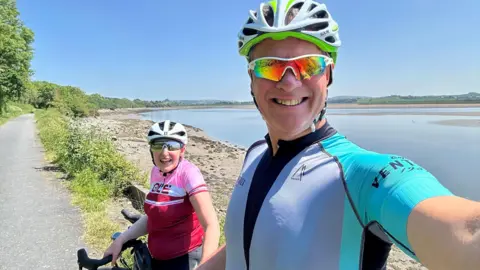 Collect/PA Real Life Sam and John take a selfie on a sunny day with their bicycles, wearing lycra and helmets. They are on a path next to large body of water