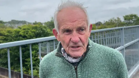 Cornwall councillor Peter Channon on a flyover bridge, with metal railings behind him. He is wearing a green knitted cardigan with a zip