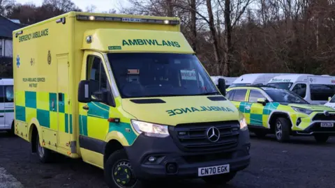 Getty Images A yellow and green ambulance vehicle, parked up with its headlights on. Behind it is a number of other ambulance service vehicles, including a green and yellow car