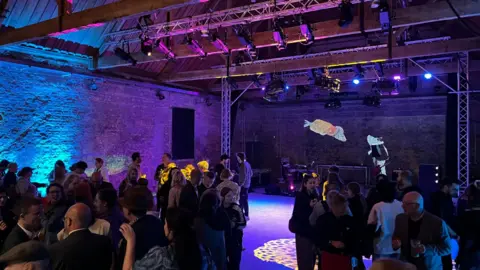 Andrew Turner/BBC The interior of Great Yarmouth's Ice House building. Brickwork is bathed in blue and purple light. There are people milling around at a launch event. A stage area has been created at one end with artworks of two fish on that wall.