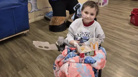 Family A young girl with short brown hair and a medical tube taped to her face sits in a wheelchair and smiles in a Build a Bear shop. A large Build a Bear teddy is stood up behind her.