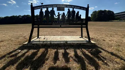 New Squids On The Block The sun streams through a metal bench made with the silhouetted figures of a group of people, from old to young. The backwards dedication reads: "To all those lost through the Covid-19 pandemic. Never Forgotten."