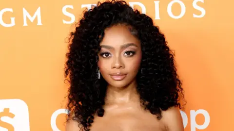 Getty Images Skai Jackson attends Variety's 2024 Power of Young Hollywood at Santa Monica Proper Hotel on August 08, 2024 in Santa Monica, California. Skai is a 22-year-old woman with dark curly hair worn loose. She has brown eyes and wears a light make-up and is styled in a sleeveless dress. She's pictured in front of an orange backdrop at the event.