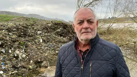 A man in a padded jacket staring into the camera lens with a pile of rubbish behind him and a river in the distance.