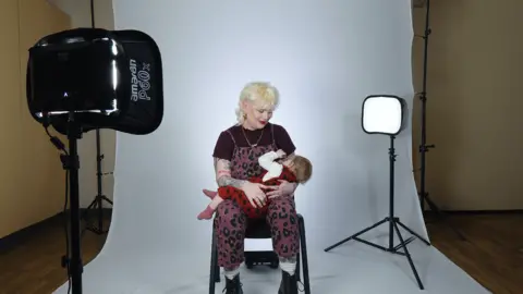 Redcar and Cleveland Council/Middlesbrough Council Emma Cooksey with her baby at the campaign photoshoot. Emma is seated and has blonde hair. She is wearing a maroon top and maroon dungarees. She has tattoos on her arms. Her child has fair hair and is wearing a red outfit. She is lying in her mum's lap. They are sitting in front of a white background with a camera on the left-hand side of the photo. A light box on top of a tripod can be seen on the right.