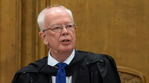 Jim Wallace is standing before the wooden walls of the Assembly Hall in Edinburgh. He is wearing a black ceremonial robe, fastened at his collar, black suit, white shirt and mid-blue tie. 