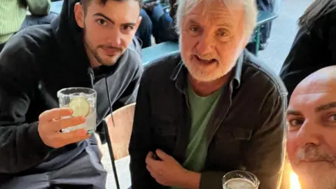 Three men sat in a pub. The one on the left is in his twenties and holding his drink aloft. The man in the middle is in his seventies and has a pint of beer next to his left arm on the table. The man on the right, who is taking the photo, is in his fifties.