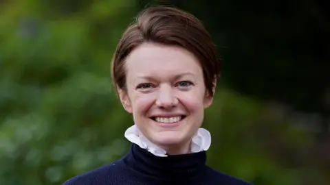 A woman with short, brown hair in a blue roll neck jumper with pink ruffled shirt showing from underneath it