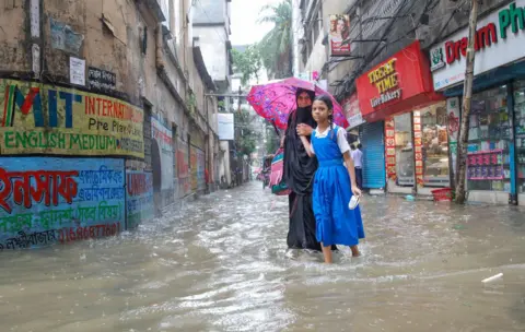 EPA A schoolgirl in a blue and white pinafore and a woman in a black dress carrying a pink umbrella wade through ankle high waters in a flooded street in Dhaka