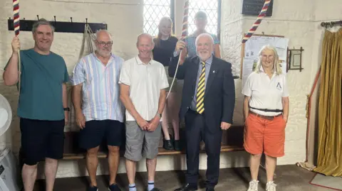 Councillor Martin Allen stood holding one of the new ropes with six bellringers at the church. He is wearing a navy blue suit with a stripey blue and white shirt, as well as a yellow and blue striped tie. 
