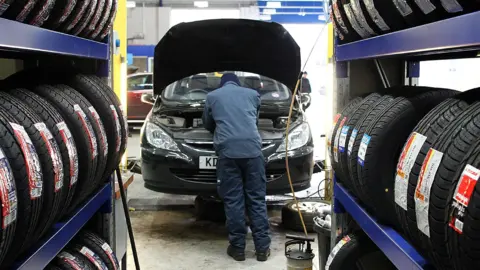 Generic image of a mechanic checking a car's engine under the bonnet inside a garage. Shelves of tyres can be seeen on both sides.