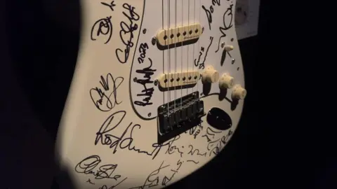 A close up of a white Fender Stratocaster guitar, covered in signatures by a series of music legends.