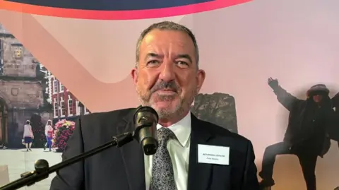 A man with short dark hair and a beard in a dark suit with a patterned tie in front of a microphone with a pink wall behind him
