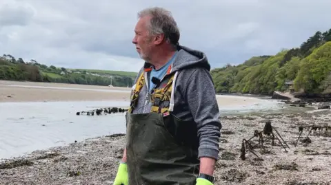 Tim Edwards is wearing waterproof waders, a hoodie, and bright gloves stands on a muddy shore beside shallow water, looking across an estuary. Around them are low racks or frames covered in seaweed, suggesting a shellfish or coastal farming area backed by green hills and trees.