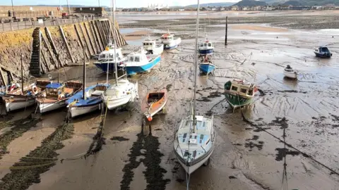 BBC Photograph of a muddy Minehead harbour at low tide. There are 14 boats dotted about the mud and chained to the harbour bed. In the background is Minehead seafront. On the left of the photo is the edge of the harbour wall.