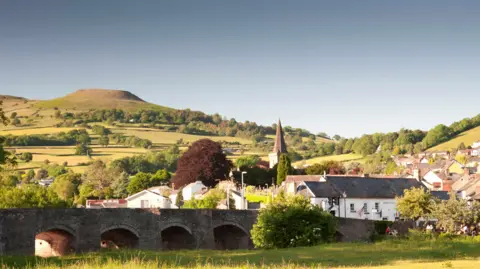 Getty Images View of Crickhowell seen from across the river with stone bridge with arches in the foreground and table mountain and green hills in the background and the village visible behind the bridge on slightly higher ground, including the spire of a church.