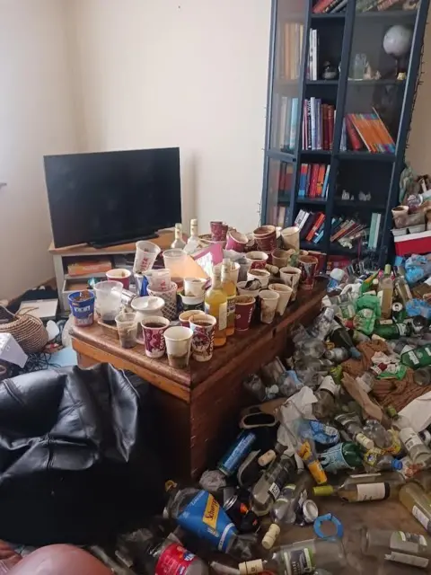 A living room with a flat screen tv, book shelf and coffee table covered with empty bottles and coffee cups.