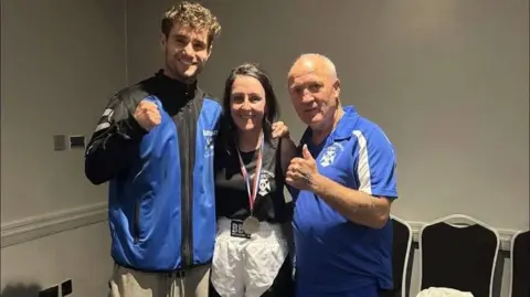 Bravehearts Amateur Boxing Club Natasha se interpone entre un joven boxeador alto en el entrenador izquierdo y mayor a la derecha, con medalla de plata alrededor de su cuello
