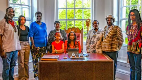 Nine people, all black, dressed in bright colours standing around a desk where a Sally Kimondo is sat. On the desk is a large book and a pen and an iPad. In the background are windows and there are trees beyond.