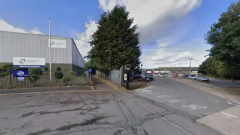 A tarmac road leads to the entrance to the warehouse. A large, grey unit stands on the left with trees standing either side of a driveway entrance.