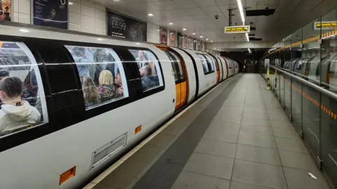 One of the new Glasgow subway trains is stationary on a platform. The train is white with orange doors and it is full of people. There is no one on the platform and the train doors are closed. Adverts can be seen behind the train.