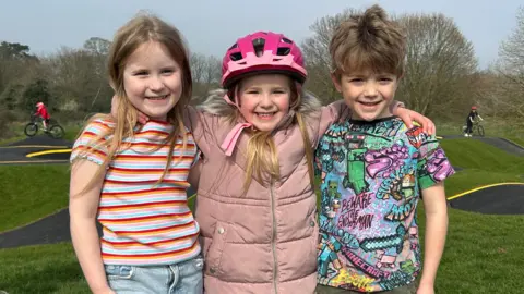 Gavin Kermack / BBC Three young children, two girls and a boy, stand next to each other with their arms around each other's shoulders in front of the pump track.