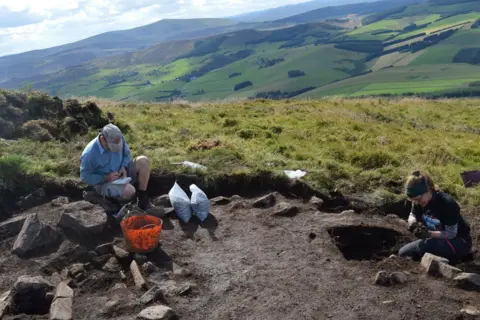 University of Aberdeen Two archaeologists sit on bare soil in an excavated area on the hill. Behind them is a landscape of rolling green, farmland.