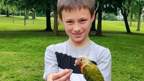Family Leo Ross, a boy with brown hair, smiles at the camera while holding a daisy in one hand. He is holding a green and white parrot-type bird which is eating the daisy.