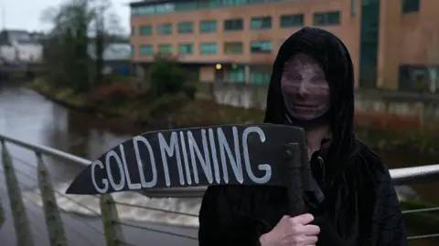 BBC Seorang pengunjuk rasa berpakaian seperti Kematian. Mereka mengenakan jubah berkerudung hitam dan memegang sabit bertuliskan penambangan emas. Wajah mereka tertutup. Sebuah sungai ada di belakang mereka. 