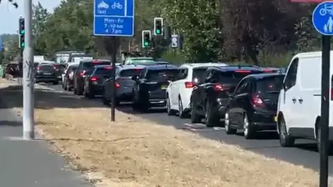 SHAM ASHRAF Daytime traffic jam on a road, showing a long queue of cars and a white van stationary near traffic lights, with blue road signs for bus and cycle lanes visible in the foreground.