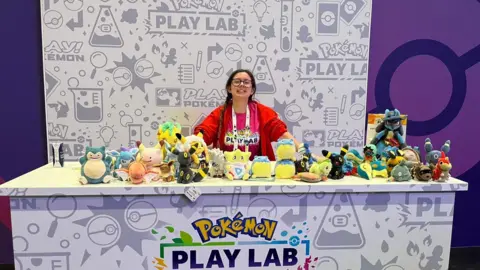 Morgan Elise A brunette girl in a red jacket is stood smiling behind a branded panel full of colourful Pokémon plushies.