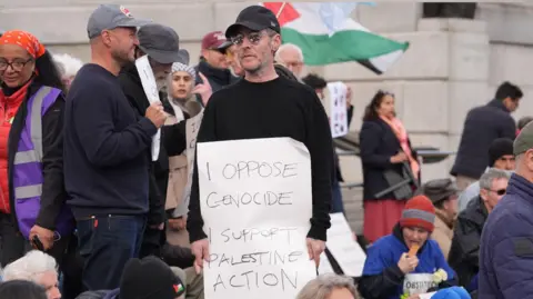 Massive Attack frontman Robert Del Naja standing at a protest at Tralfalgar Square holding a sign that reads: "I oppose genocide I support Palestine Action." He is wearing a black baseball cap, sunglasses and a black jumper