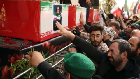 The image depicts a solemn funeral ceremony in Iran. Several red coffins, each draped with the national flag of Iran, are prominently displayed. The flag’s green, white, and red tricolor with Persian script is visible, emphasizing the national significance of the event. One of the coffins bears a portrait of a man in military uniform—likely honouring someone viewed as a martyr or key figure. Mourners are gathered closely, some reaching out to touch the coffins, while others hold green sprigs