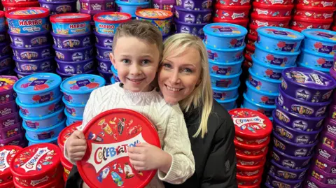 Teddy and Laura in the warehouse with the sweet tubs. Teddy holds a tub of Celebrations. His mum rests her head on his shoulder. She has long blonde hair and wears a black coat.