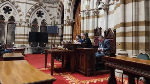 Nadia Lincoln/LDRS A number of people in a meeting, with three people sitting on large wooden chairs and the woman in the middle holding a piece of paper. There is a large red carpet on the floor, and large columns all around the room. There are also a number of TV screens.