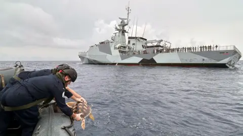 PA Media The crew of a Royal Navy warship are pictured at the release of six rare turtles. In the background you can see the warship and near to the camera you can see a smaller boat with the turtle being released.