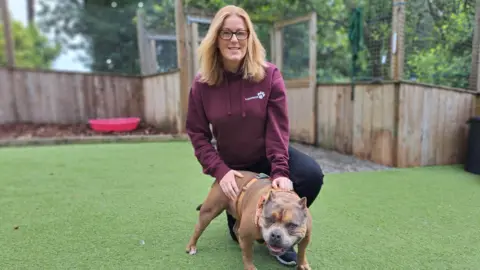Vanessa wearing a burgundy hoodie with the Hope Rescue name on it, crouched by a tan dog with cropped ears. Vanessa has long blonde hair and is wearing glasses.