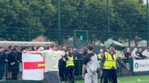 BBC Football fans stood on a pitch. Some are stood behind a gate. There is a bin in the air. There are trees in the background. 