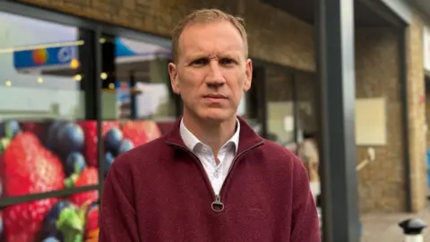 Dónal McAnallen has short brown hair and is wearing a maroon quarter-zip over the top of a white shirt. In the blurred background is a shopfront with sandstone brick and windows with advertising for strawberries and blueberries.