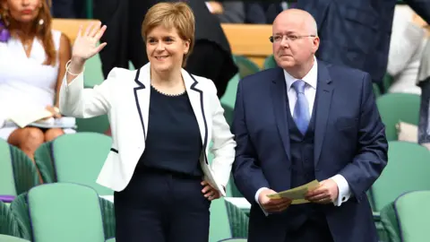 Getty Images Sturgeon and Murrell at Wimbledon in 2016. They are standing among rows of green seats; She is wearing a white jacket over a dark outfit and raises a hand in greeting, while he wears a dark suit and holds a paper programme.