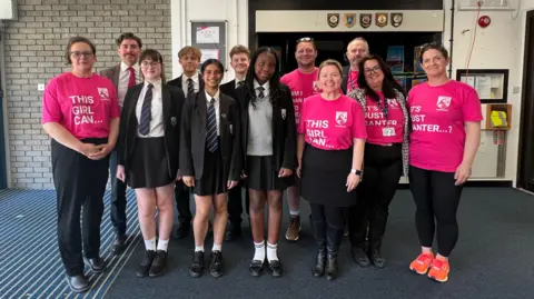 Photo of teachers and students standing in two rows. There are four female teachers wearing pink T-shirts with slogans on including as 'this girl can and 'it's just banter...?'. There are two male teachers with ink T-shits on and one male teacher with a brown blazer and pink tie. There are three female students in black blazers, white shirts and black skirts with a blue striped tie. There are two male students also in a black blazer, white shirt and blue tie.