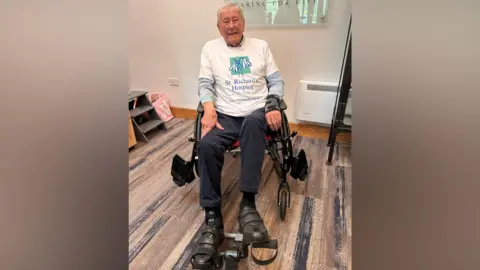 St Richard’s Hospice The image shows an Ken Johnson sitting on a chair using a small stationary pedal exerciser placed on the floor in front of them. The person is wearing a white T-shirt with a printed logo and text that reads “St. Richard’s Hospice – Caring for Life.” A supportive walking frame is positioned in front of the exerciser, and the person is wearing supportive footwear or braces on both feet.