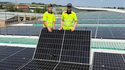 Two men in yellow high-vis jackets stand among dozens of solar panels on the roof of a building